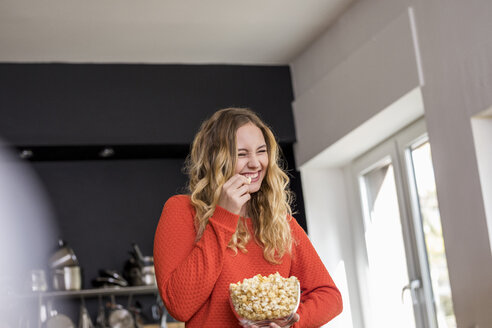 Portrait of giggling young woman with bowl of popcorn in the kitchen - FMKF04653