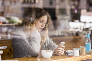 Porträt einer traurigen jungen Frau in einem Café, die auf ihr Mobiltelefon schaut - FMKF04658