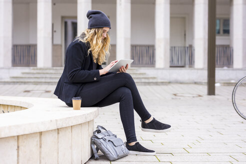 Junge Frau mit Kaffee zum Mitnehmen sitzt an einer Wand und benutzt ein Tablet - FMKF04668