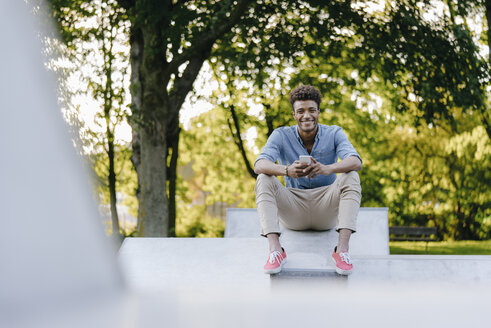 Lächelnder junger Mann mit Handy im Skatepark sitzend - KNSF03184