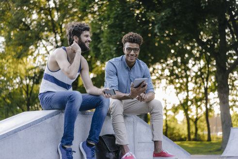 Zwei glückliche Freunde sitzen in einem Skatepark mit mobilen Geräten - KNSF03194