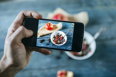 Männerhand, die mit einem Smartphone eine Schüssel mit einem Dessert aus Mandeln, Granatapfel und Schokolade fotografiert - KIJF01795