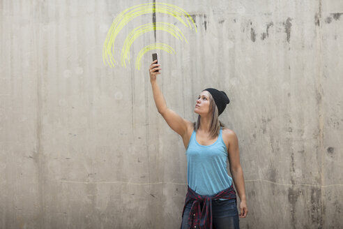 Frau hält Telefon auf der Suche nach Signal mit wifi Zeichen in gelber Kreide auf Betonwand gezeichnet - ZEF14874