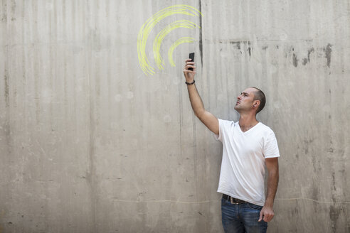 Mann hält Telefon auf der Suche nach Signal mit wifi Zeichen in gelber Kreide auf Betonwand gezeichnet - ZEF14875