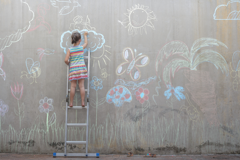 Girl standing on ladder drawing colourful pictures with chalk on a concrete wall stock photo