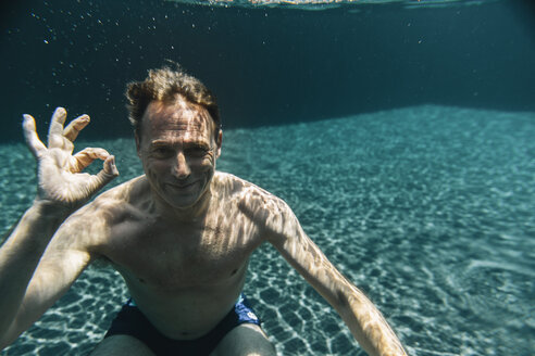 Porträt eines lächelnden Mannes unter Wasser in einem Schwimmbad, der ein Ok-Zeichen macht - MFF04230