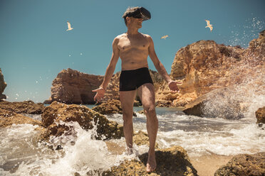 Mature man wearing VR glasses between rocks on beach - MFF04264