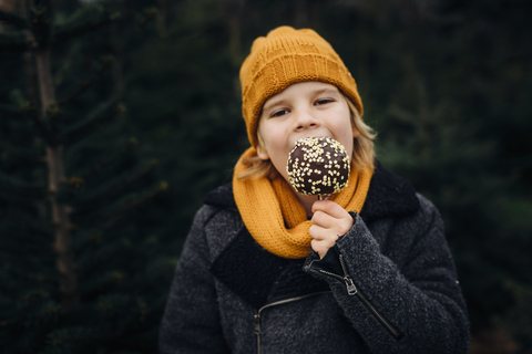 Kleiner Junge steht vor einer Tanne und isst einen in Schokolade getauchten Apfel, lizenzfreies Stockfoto