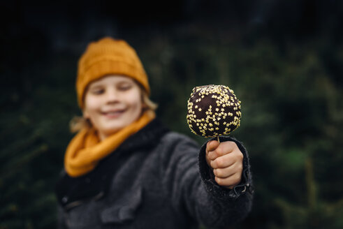 Little boy standing in front of fir trees holding chocolate dipped apple - MJF02242