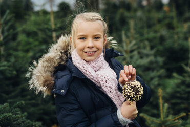 Little girl standing in front of fir trees holding chocolate dipped apple - MJF02244