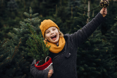 Fröhlicher Junge bereitet sich auf Weihnachten vor, hält einen Topfbaum und isst einen in Schokolade getauchten Apfel - MJF02246