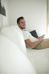 Lächelnder junger Mann, der sich zu Hause mit seinem Smartphone entspannt - RAEF01946