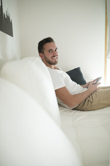 Lächelnder junger Mann, der sich zu Hause mit seinem Smartphone entspannt - RAEF01946