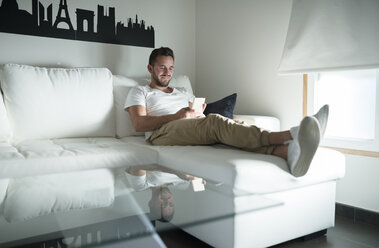 Smiling young man relaxing at home with his smartphone - RAEF01947