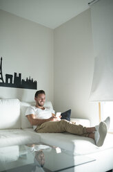 Smiling young man relaxing at home with his smartphone - RAEF01948