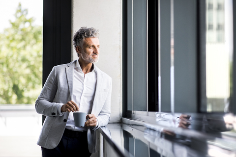 Lächelnder reifer Geschäftsmann mit Kaffeetasse, der aus dem Fenster schaut, lizenzfreies Stockfoto