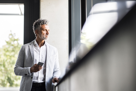 Reifer Geschäftsmann mit Handy schaut aus dem Fenster, lizenzfreies Stockfoto