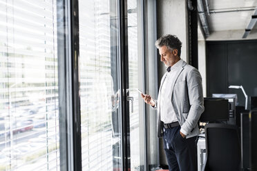 Älterer Geschäftsmann steht am Fenster im Büro und hält Handy - HAPF02557