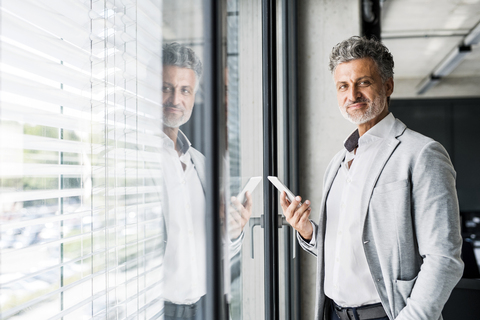 Porträt eines lächelnden reifen Geschäftsmannes am Fenster im Büro, der ein Mobiltelefon hält, lizenzfreies Stockfoto