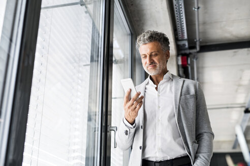Smiling mature businessman standing at the window in office holding cell phone - HAPF02559