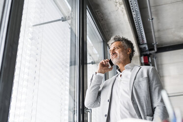 Smiling mature businessman on cell phone at the window in office - HAPF02560