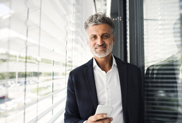 Portrait of smiling mature businessman holding cell phone at outside sunblind - HAPF02566