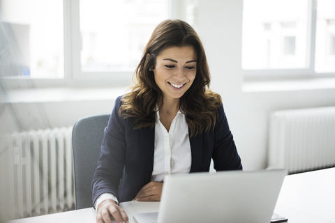 Porträt einer lächelnden Geschäftsfrau, die an einem Schreibtisch im Büro sitzt und an einem Laptop arbeitet, lizenzfreies Stockfoto