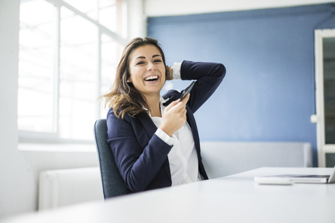 Portrait of laughing businesswoman with cell phone sitting at desk in the office stock photo