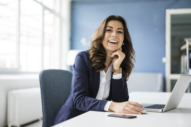 Portrait of laughing businesswoman sitting at desk in the office - MOEF00457