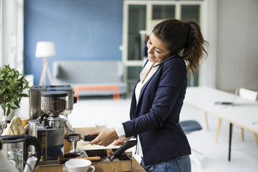 Lächelnde Geschäftsfrau am Telefon bei der Zubereitung von Espresso mit Espressomaschine in einem Loft - MOEF00466