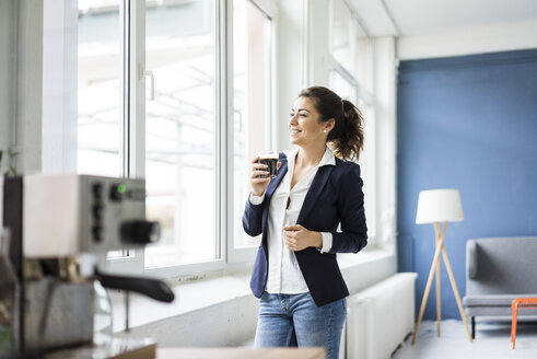 Glückliche Geschäftsfrau mit einem Glas Kaffee in einem Loft und Blick aus dem Fenster - MOEF00468