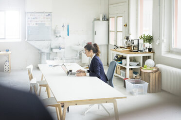Lächelnde Geschäftsfrau sitzt am Schreibtisch in einem Loft und arbeitet am Laptop - MOEF00470