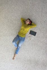 Lächelnde junge Frau, die mit Kopfhörern und Tablet auf dem Boden liegt und Musik hört, Ansicht von oben - MOEF00506