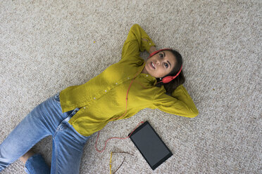Porträt einer jungen Frau, die mit Kopfhörern und Tablet auf dem Boden liegt und Musik hört, Ansicht von oben - MOEF00507