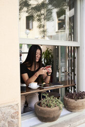 Lächelnde asiatische Frau, die in einem Café neben dem Fenster mit ihrem Telefon plaudert - IGGF00296