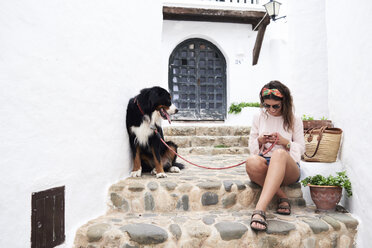 Smiley brunette girl chatting with phone while holding dog lead of her bernese dog in summer - IGGF00308