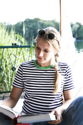Young woman reading book at a lake next to sailing boat - FKF02792