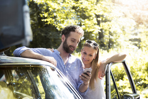 Smiling young couple with cell phone at car in forest - FKF02793