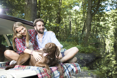 Smiling young couple with dog sitting in car at a brook in forest stock photo