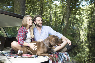 Smiling young couple with dog sitting in car at a brook in forest - FKF02806