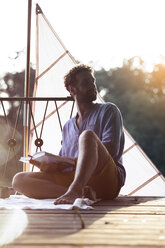 Young man with book sitting on a jetty next to sailing boat - FKF02822