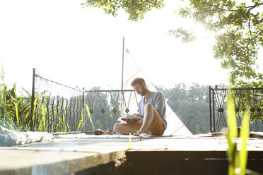 Young man reading book on a jetty next to sailing boat - FKF02835