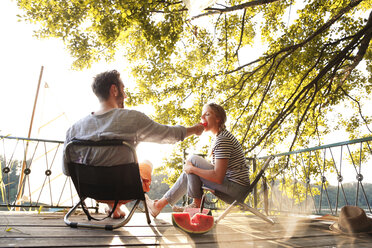 Young couple sitting on a jetty at a lake eating watermelon - FKF02845