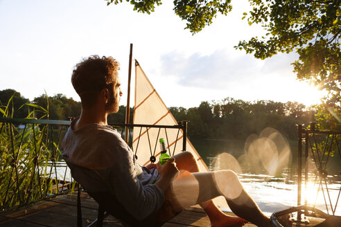 Young man with beer bottle sitting on a jetty next to sailing boat - FKF02846