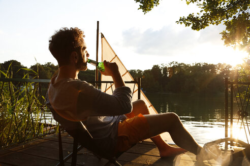 Young man with drinking a beer on a jetty next to sailing boat - FKF02847