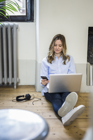 Frau sitzt zu Hause auf dem Boden und benutzt Handy und Laptop, lizenzfreies Stockfoto