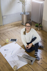 Smiling woman sitting on the floor with blueprint,cell phone and laptop - GIOF03622