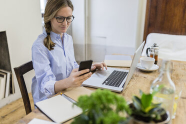 Woman using cell phone and laptop on wooden desk at home - GIOF03634