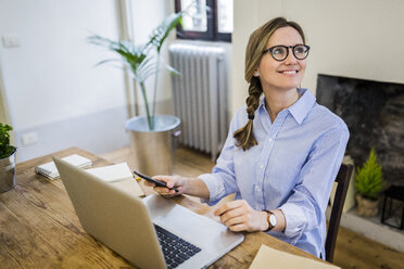 Smiling woman sitting at wooden desk at home looking sideways - GIOF03635