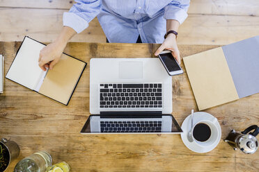 Top view of woman at wooden desk with notebook, cell phone and laptop - GIOF03636